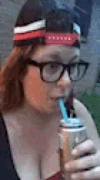 a woman wearing glasses and a hat is drinking from a can .