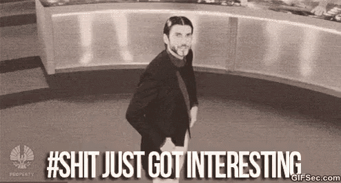 a man in a suit and tie is standing in a room with the words `` shit just got interesting '' written above him .