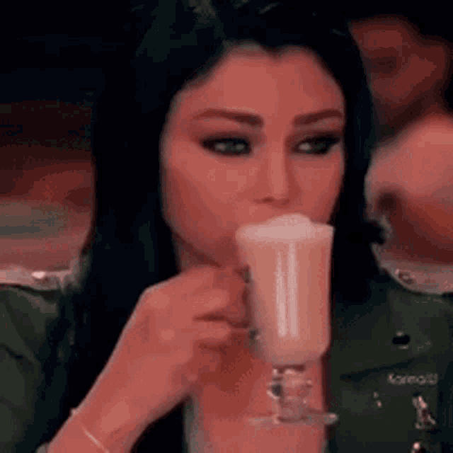 a woman is drinking a milkshake from a glass while wearing a green jacket .