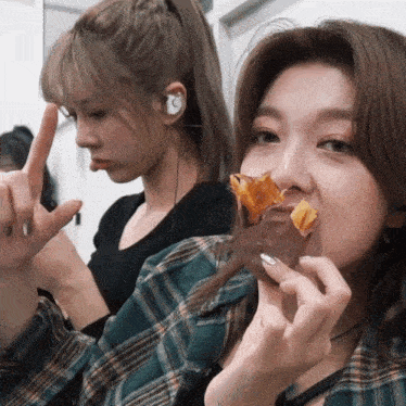 a woman eating a piece of food next to another woman wearing ear buds