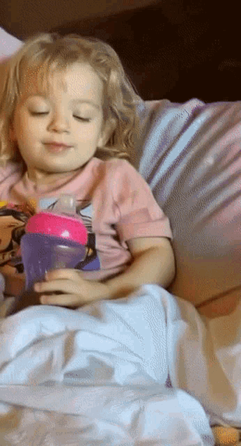 a little girl is sitting on a bed holding a purple cup