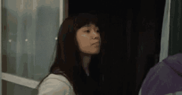 a woman with long black hair is standing in front of a window and looking out .