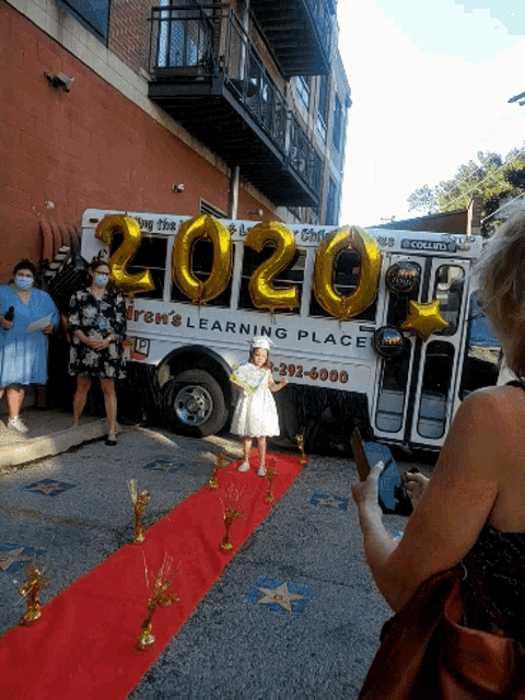 a little girl is standing on a red carpet in front of a bus that says 2020