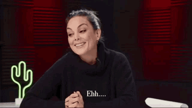 a woman in a black sweater is smiling and sitting in front of a neon cactus .