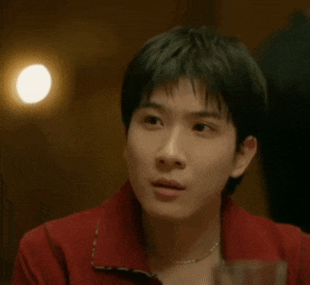 a young man in a red shirt is sitting at a table with a glass of wine and making a surprised face .