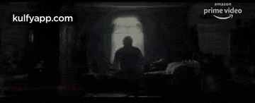 a man is sitting in front of a window in a dark room with a silhouette of a person behind him .