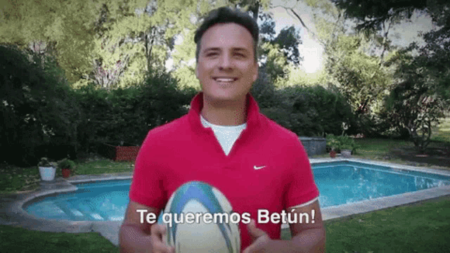 a man in a red shirt holds a rugby ball in front of a pool and says " te queremos betun "