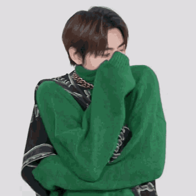 a young man is wearing a green sweater and covering his face with his hands .