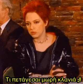a woman with red hair is sitting at a table in front of a brick wall with a man in a suit in the background .