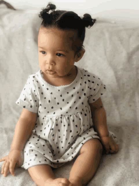 a baby in a white dress with black polka dots sits on a bed