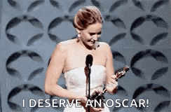 a woman in a white dress is holding an oscar trophy and says `` i deserve an oscar ! ''