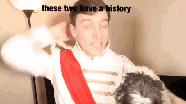 a man in a costume with a red sash is holding a dog with the caption " these two have a history " above him