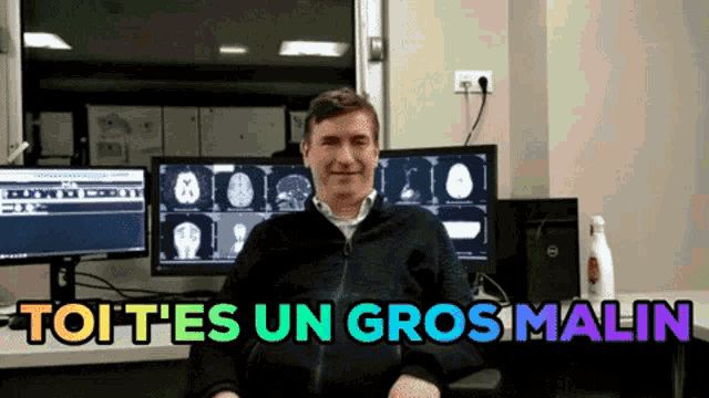 a man sitting in front of two computer monitors with the words " toutes un gros malin " written above him