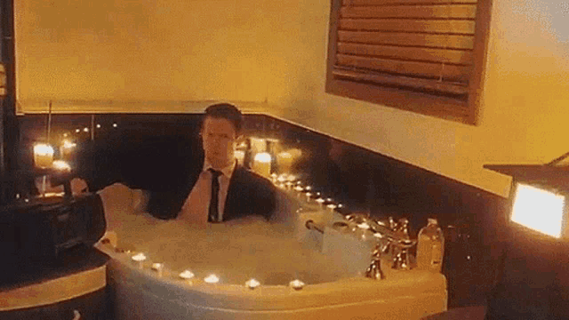 a man in a suit and tie is taking a bath in a jacuzzi surrounded by candles .