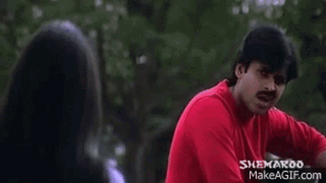 a man in a red shirt and mustache is talking to a woman in a park .