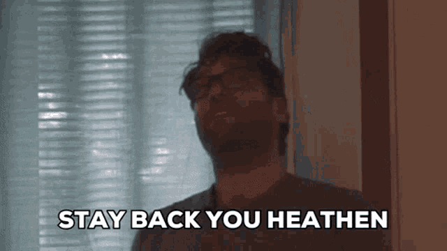 a man with glasses says " stay back you heathen " in front of a window