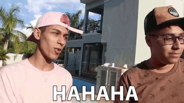 a man wearing glasses and a baseball cap laughs next to another man wearing glasses and a pink shirt