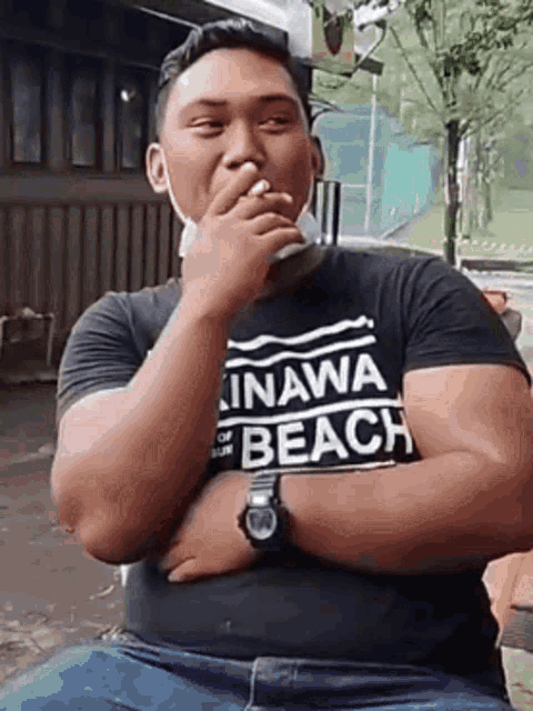 a man wearing a black shirt with the word beach on it is smoking a cigarette while sitting on a bench .