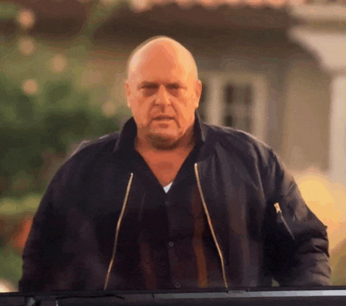a bald man in a black jacket stands in front of a building