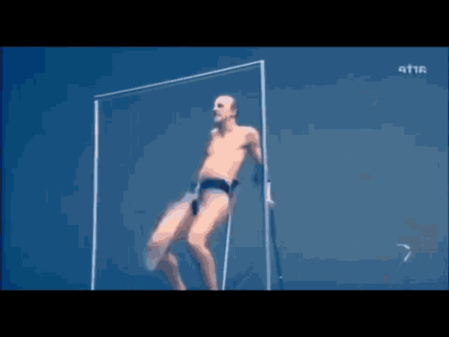 a man without a shirt is standing in a glass box with his legs crossed .