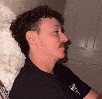 a man with curly hair and a mustache is wearing a black shirt with a skull and crossbones embroidered on it