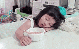 a little girl is sleeping on a table with a bowl of cereal in front of her .