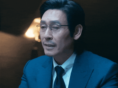 a man wearing glasses and a blue suit is sitting in a dark room
