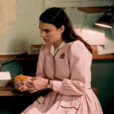 a woman in a pink dress is sitting at a desk eating a sandwich