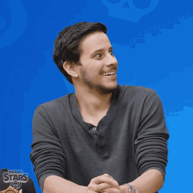 a man in a grey shirt is smiling in front of a blue background that says brawl stars