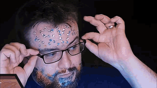 a man with glasses and glitter on his face has the number 77 on the bottom right
