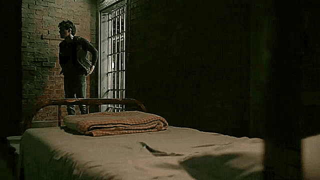 a man is sitting on a bed in a dark room looking out of a window .