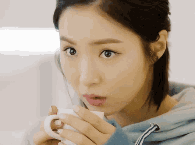 a woman in a blue sweatshirt is drinking from a white mug