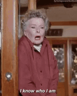 a woman in a red shirt is standing in front of a door and says `` i know who i am '' .
