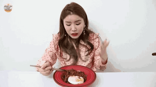 a woman in a pink polka dot shirt is sitting at a table eating a bowl of food with chopsticks .