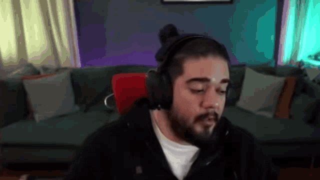 a man with a beard wearing headphones is sitting in front of a couch in a living room .