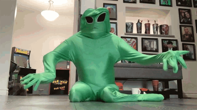 a person in a green alien costume is sitting on the floor in front of a pac man arcade machine