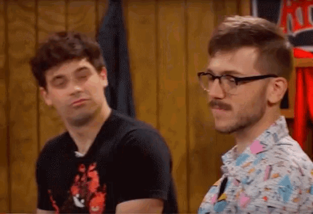 two men are sitting next to each other in front of a wooden wall . one of the men has glasses on .