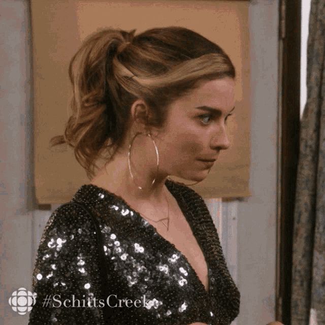 a woman wearing hoop earrings and a sequined top with #schitts creek on the bottom