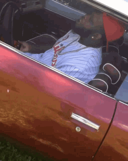 a man in a baseball uniform is sitting in the driver 's seat of a red car