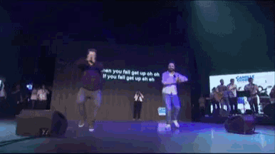 two men are dancing on a stage in front of a screen that says " can you fall get up oh oh "