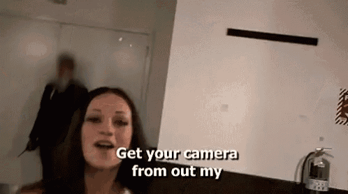 a woman is standing in a room talking to a man and asking him to get his camera from out of her .