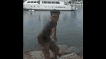 a boy is standing on a rock in front of a body of water with a boat in the background .