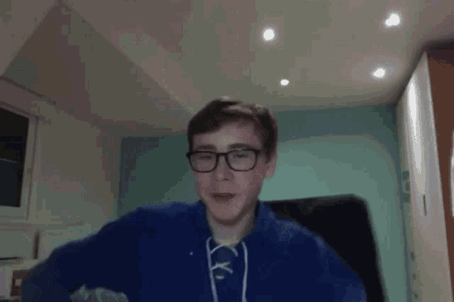 a young man wearing glasses and a blue sweater is sitting in a chair in a room .