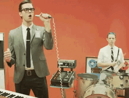 a man in a suit is singing into a microphone while a drummer plays drums