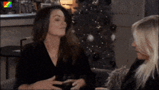 two women are sitting in front of a christmas tree drinking wine