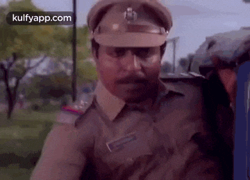 a man in a police uniform with a mustache is standing in a park with trees in the background .