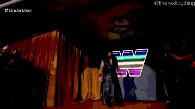 a man in a black coat is walking down a stage with the word undertaker on the bottom left