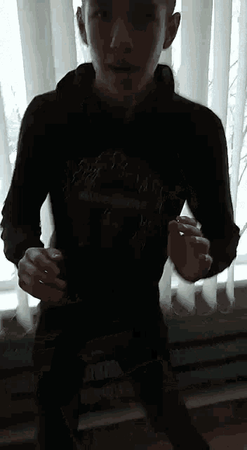 a young man in a black hoodie is standing in front of a window with blinds .