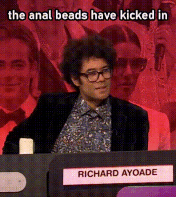 richard ayoade is sitting at a table with a sign that says richard ayoade