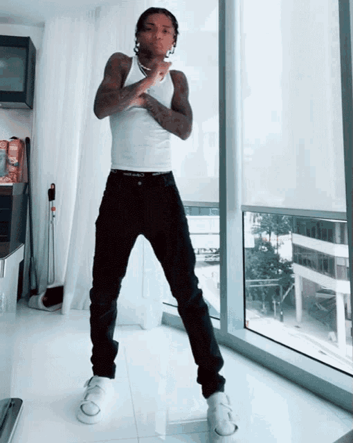 a man in a white tank top and black jeans standing in front of a window